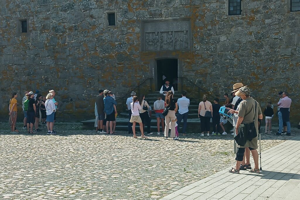 På trappan till den medeltida borgen står en guide i tidstypiska kläder med en grupp turister. Lite vid sidan av står ett par med en digital guide i surfplattan. Kvinnan, i khakifärgad T-shirts och shorts i samma färg har krycka och knästöd.