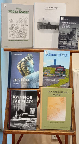 Sju böcker och häften i olika storlekar och färger står uppställda på ett trästafli.