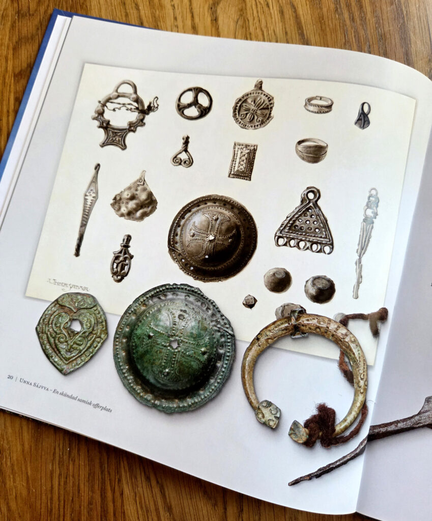 En uppslagen bok där en sida visar fotografier av flera metallföremål såsom broscher, hängen och knappar. På boksidan ligger också några riktiga föremål placerade ovanpå, bland annat en rund bronsplatta och ett metallspänne.