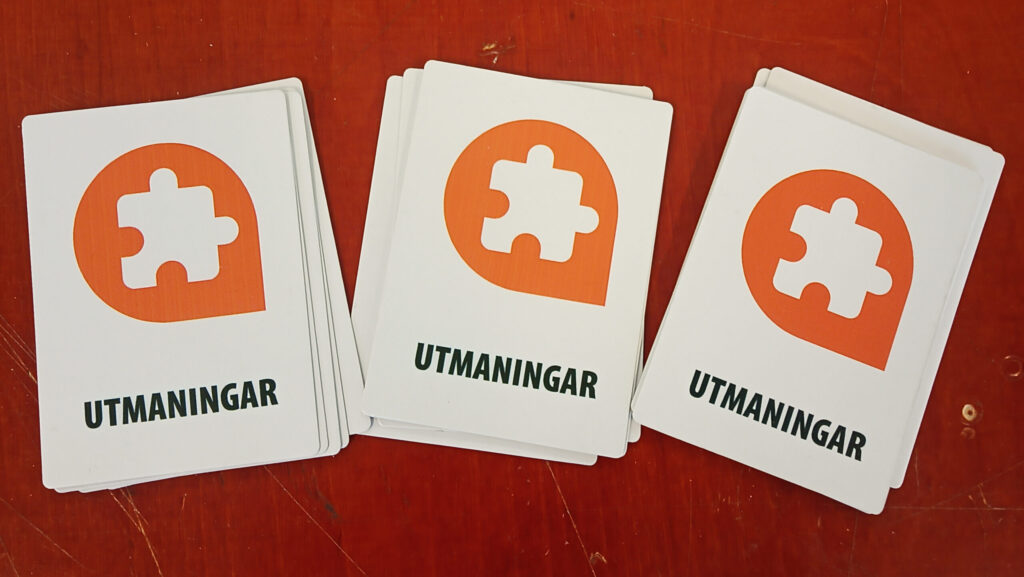 En kortlek, där tre högar med kort ligger med baksidorna uppåt. På baksidorna finns en bild av en pusselbit innesluten i det övre vänstra av Riksantikvarieämbetets orangea logotyp. På varje kort finns också texten "Utmaningar".
