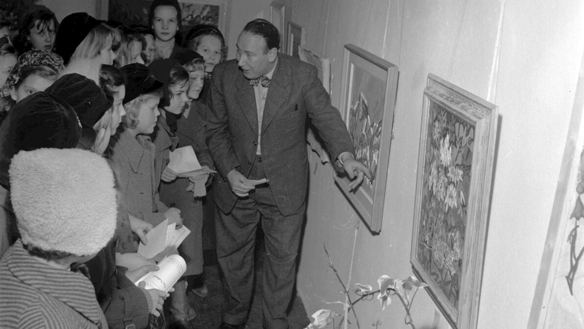 Svartvit bild från en konsthall där barn tittar på målningar som presenteras av en man i kostym