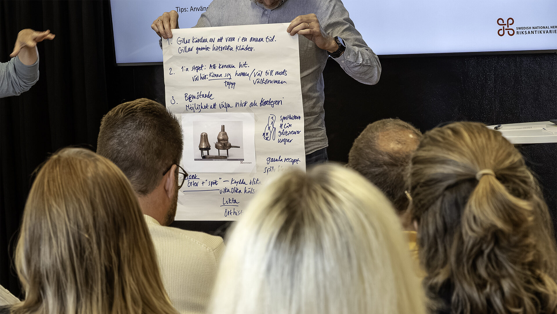 Ett papper med förslag på en utställning i text och bilder hålls upp på en scen inför publik.