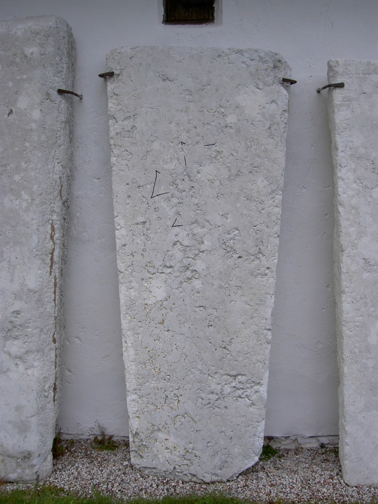Bilden visar en trapetsoid gravhäll med ett kors rest mot en vägg.