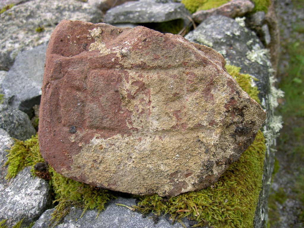 Bilden visar ett runstensfragment på en stenmur med mycket bruk på ristningsytan