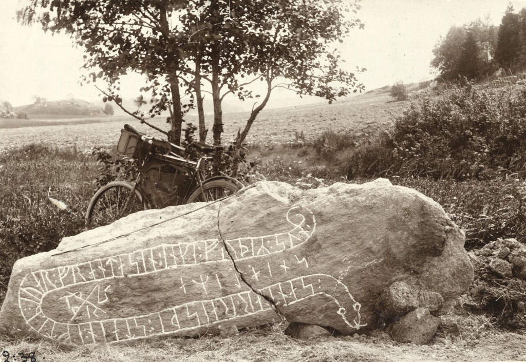 Äldre foto föreställande en liggande runsten med en cykel och ett träd i bakgrunden