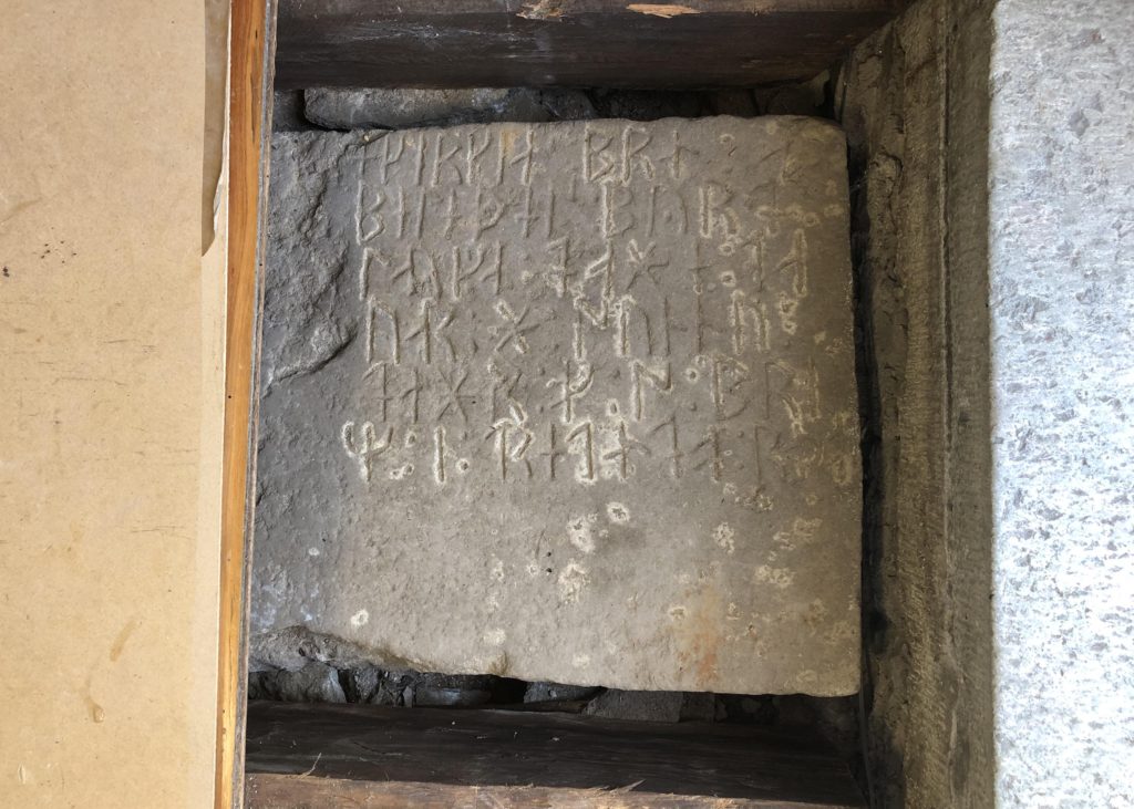 Bilden visar en fyrkantig sten med runor i ett uppsågat hål under ett golv
