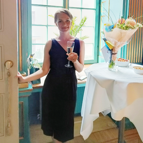 Emma Jansson med champagneglas i handen under sin disputation. Blommor på bordet bredvid henne.