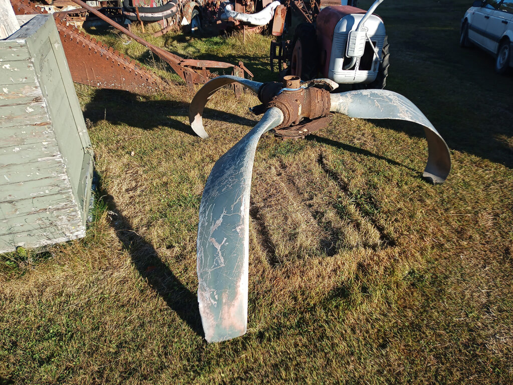 En något repad och tillbucklad flygplanspropeller på en gräsmatta. Omkring den skymtar diverse gamla maskiner.