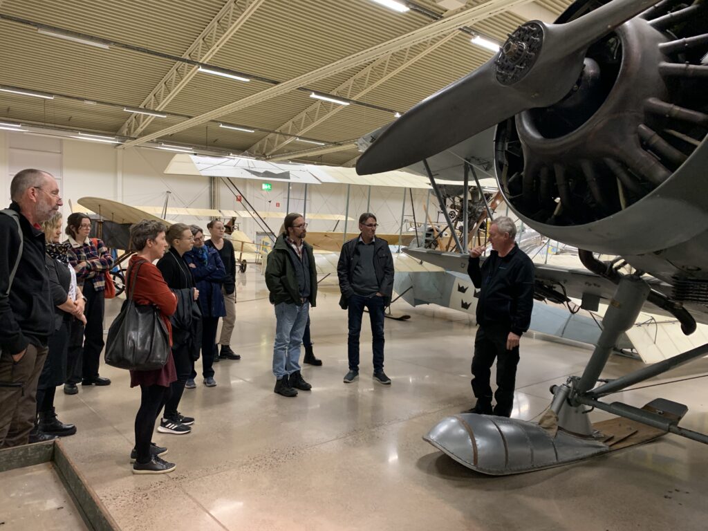 Ett tiotal människor står runt ett propellerflygplan som är utställt på ett museum. En man pekar och visar.