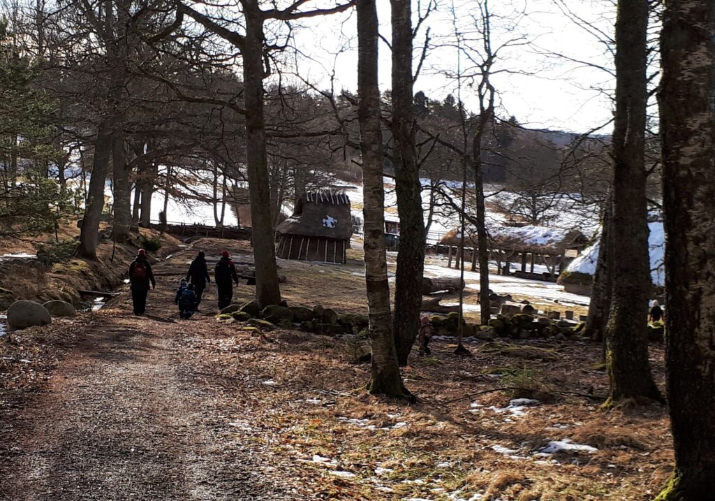 Barn och vuxna går på en väg genom en vintrig lövskog. I bakgrunden syns byggnader med stråtak.