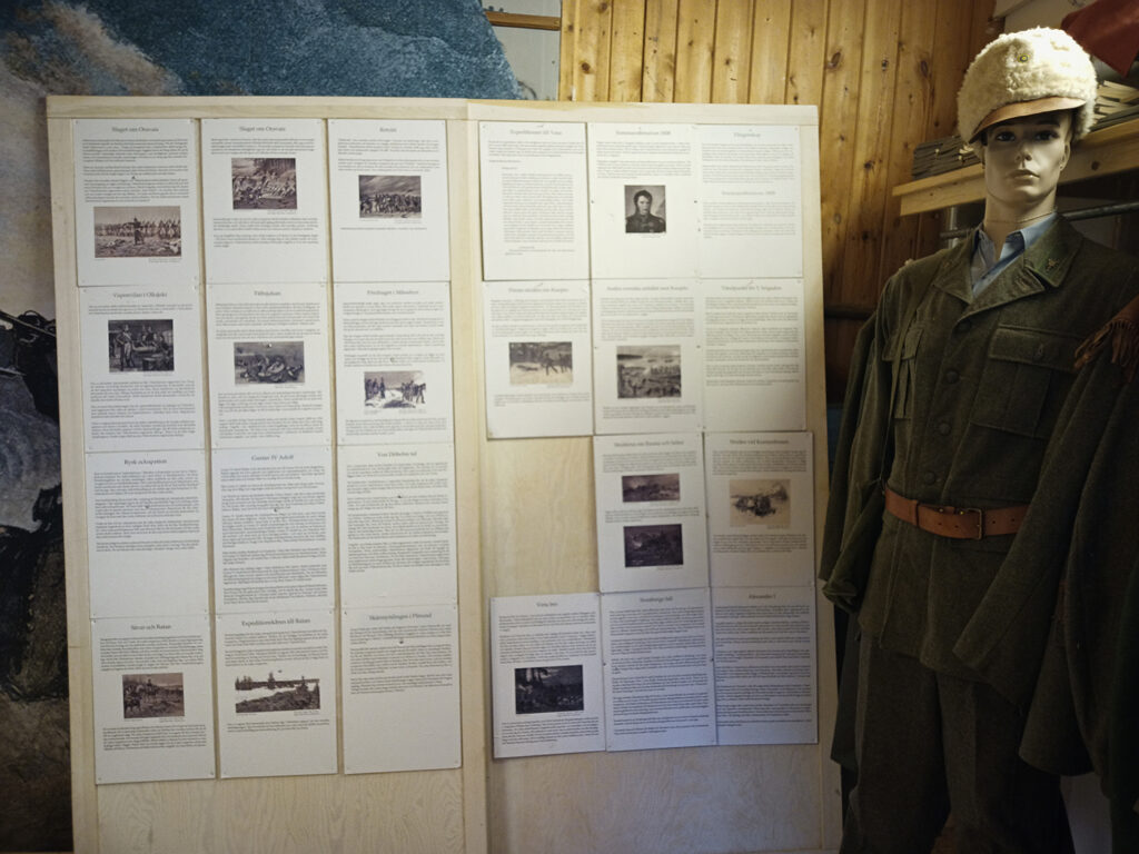 Inne i en del av Kalixlinjens museum. I bild syns en skärmvägg täckt av A4-ark med texter och bilder. Till höger står en skyltdocka iförd en äldre militäruniform med vit pälsmössa.
