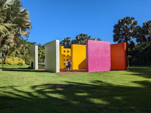 Tre personer går omkring vid sju väggar i olika färger, två gula, två vita, en rosa och en röd. Grönt gräs och träd runt omkring.