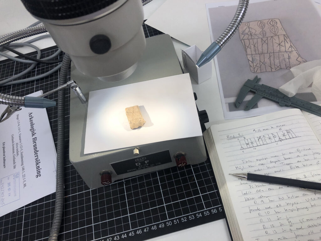 Bilden visar ett runbleck under ett mikroskop omgivet av olika föremål som anteckningsbok, skjutmått, en kalkering med mera