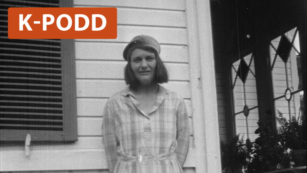 Kvinna i rutig klänning och hatt står mot en husvägg och tittar in i kameran. 1930-tal.