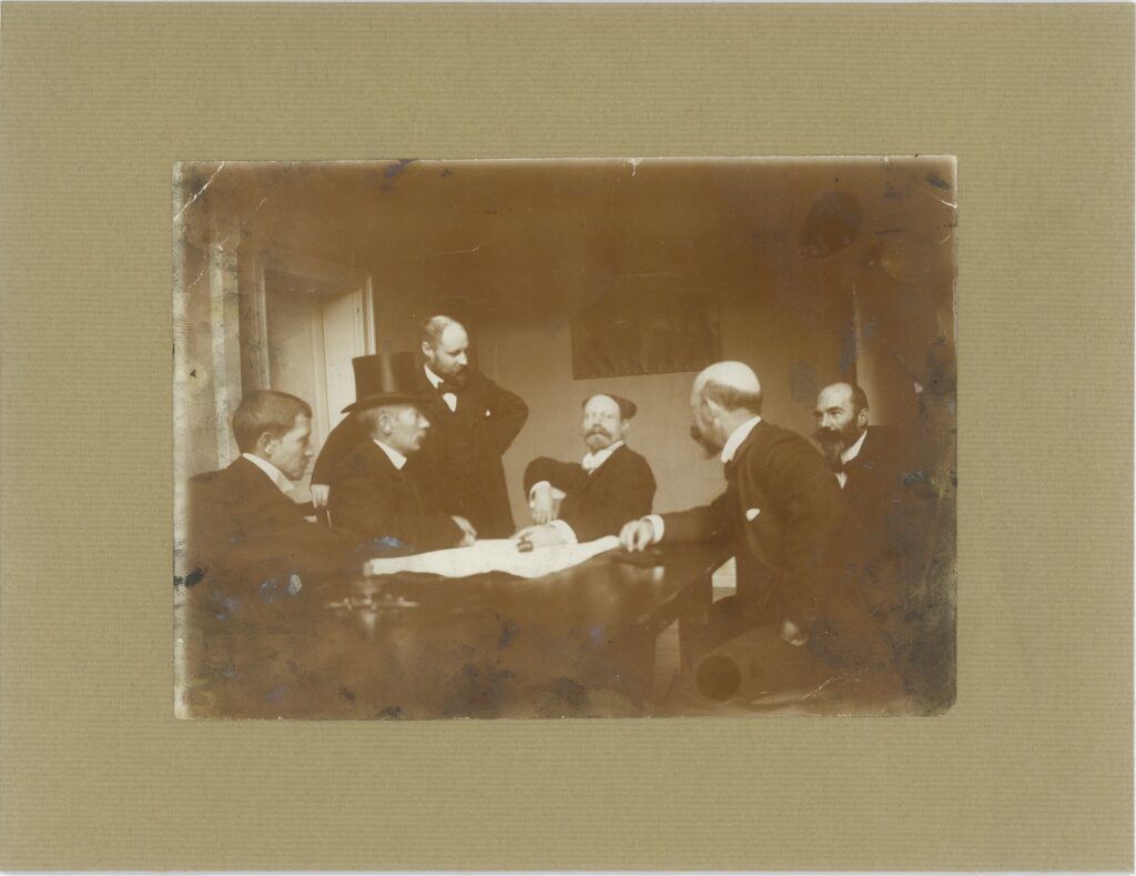 Svartvitt foto med fem män runt ett brunt stort bord. Fyra av dem sitter, en står och en har hög hatt. En man håller en ordförandeklubba.