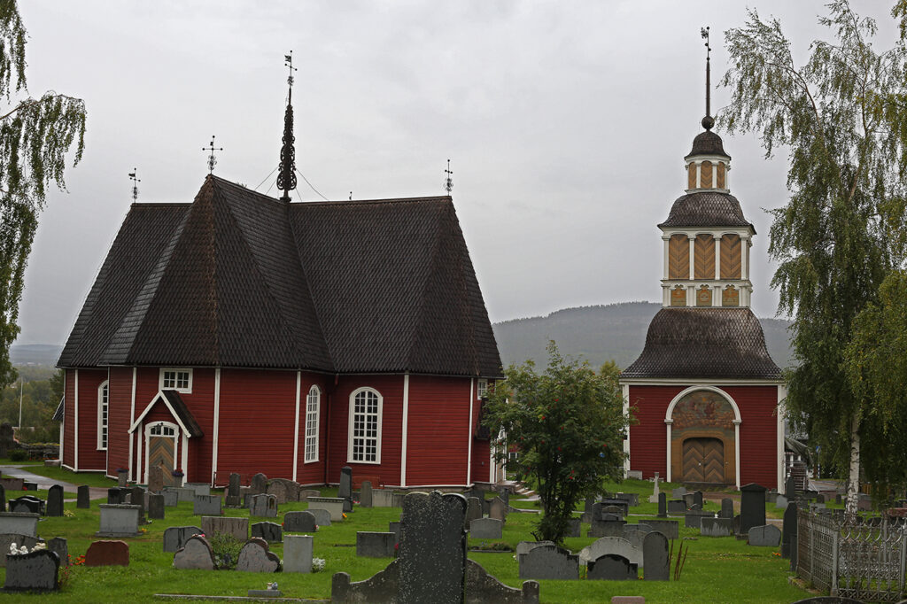 Röd korskyrka med sticktak och klockstapel av bottnisk typ. Det är sensommar och mulet, men björkarna är fortfarande gröna. Framför kyrkan syns gravstenar och kors på kyrkogården. I bakgrunden syns berget Aavasaksa på finska sidan Torneäven.