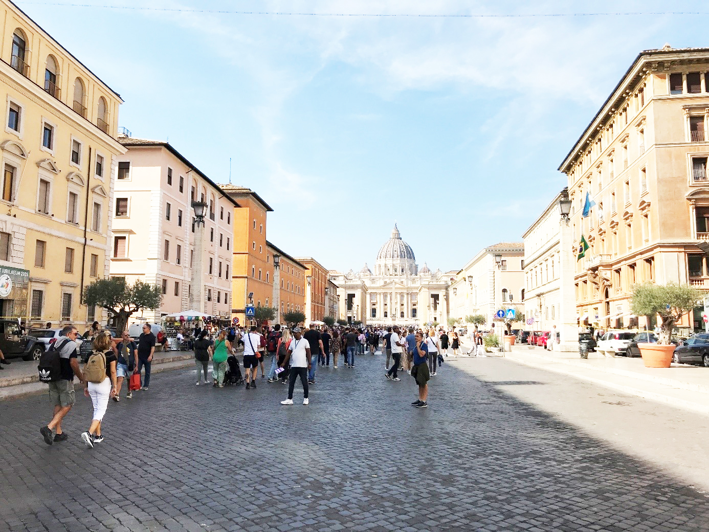 Bild av en bred gata med fotgängare på Via della Conciliazone i Rom. Peterskyrkans kupoltak i bakgrunden.