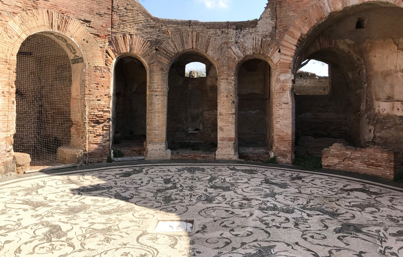 Bild av det runda rummet med golvmosaik i badhuset Terme dei Sette sapienti i Ostia Antica, Rom. Valvbågar i tegel, arkader runt det dekorerade golvet av mosaik.
