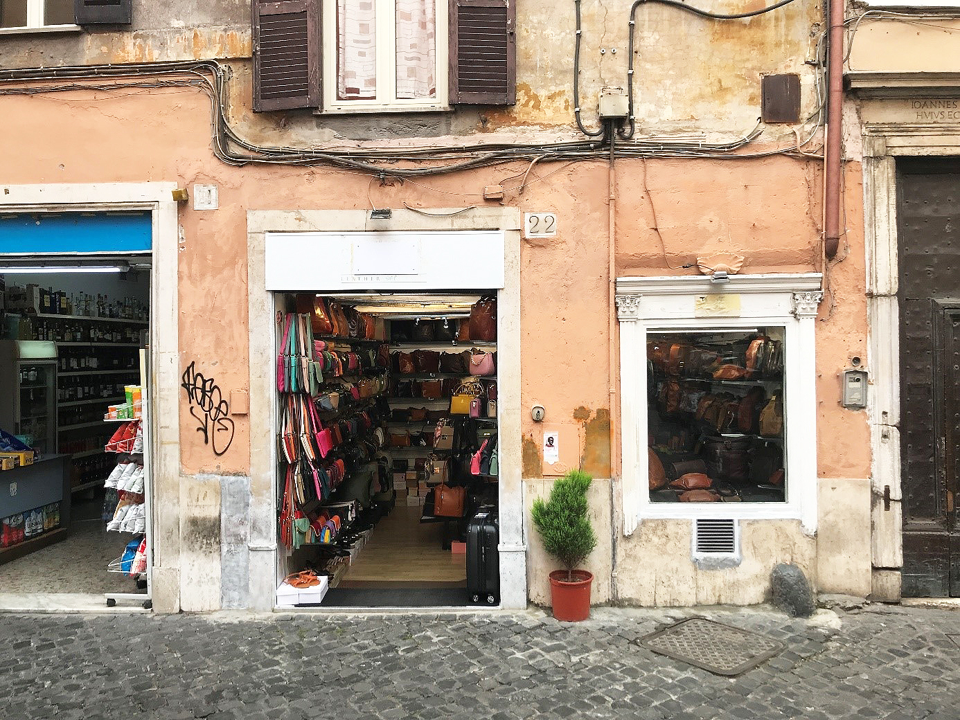 Bild av en butikfasad vid en stenlagd gata i stadsdelen Trastevere i Rom. Skyltfönster, öppna dörrar, hyllor med varor, handväskor, påsar.