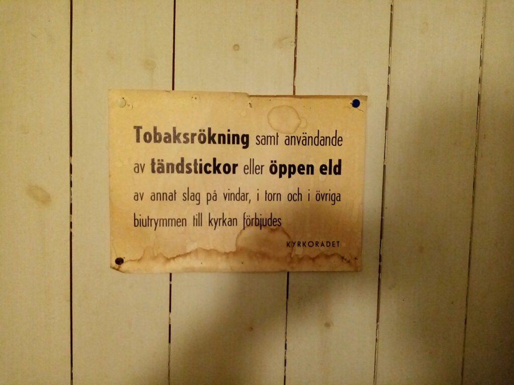 En gulnad och fuktskadad skylt med följande text: "Tobaksrökning samt användande av tändstickor eller öppen eld av annat slag på vindar, i torn och i övriga biutrymmen till kyrkan förbjudes. Kyrkorådet"
