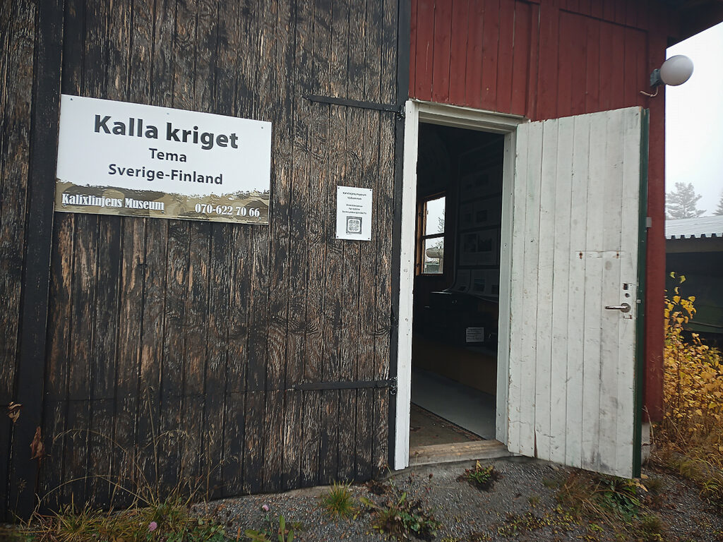 En träbyggnad med stora garagedörrar, och en mindre dörr som står öppen. På den ena garagedörren sitter en skylt med texten "Kalla kriget, Tema Sverige-Finland" och en QR-kod.