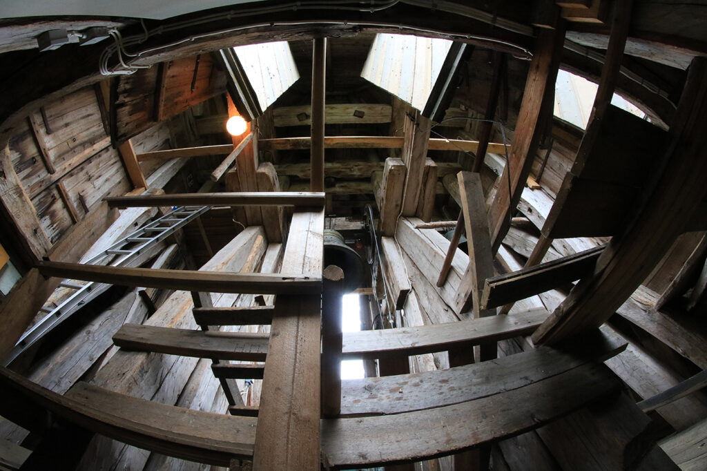 Träkonstruktion och klocka inne i klockstapeln. Fotot är taget nerifrån och upp med fisheyeobjektiv, vilket ger en känsla av höjd och svindel. Till vänster syns en stege upp till klockan.