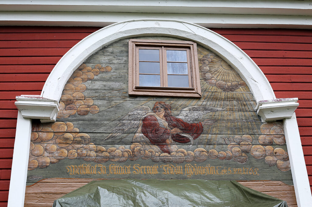 En målad panel ovanför den södra ingången till klockstapeln. En ängel med vingar och en vit klädnad med vida röda ärmar står bland molnen, och håller en skära i sin högra hand, I högra hörnet finns en strålande sol. Under ängeln och molnen finns själva bibeltexten, skriven i frakturstil på finska.