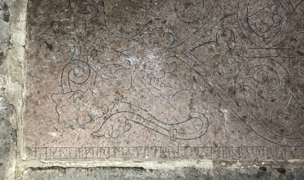 Bilden visar en del av gravhäll av röd kalksten med ornament och en rad med runor längs den nedre kanten