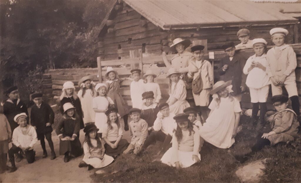 En grupp barn sitter och står framför en timmerbyggnad. En kvinna i hatt står i mitten.