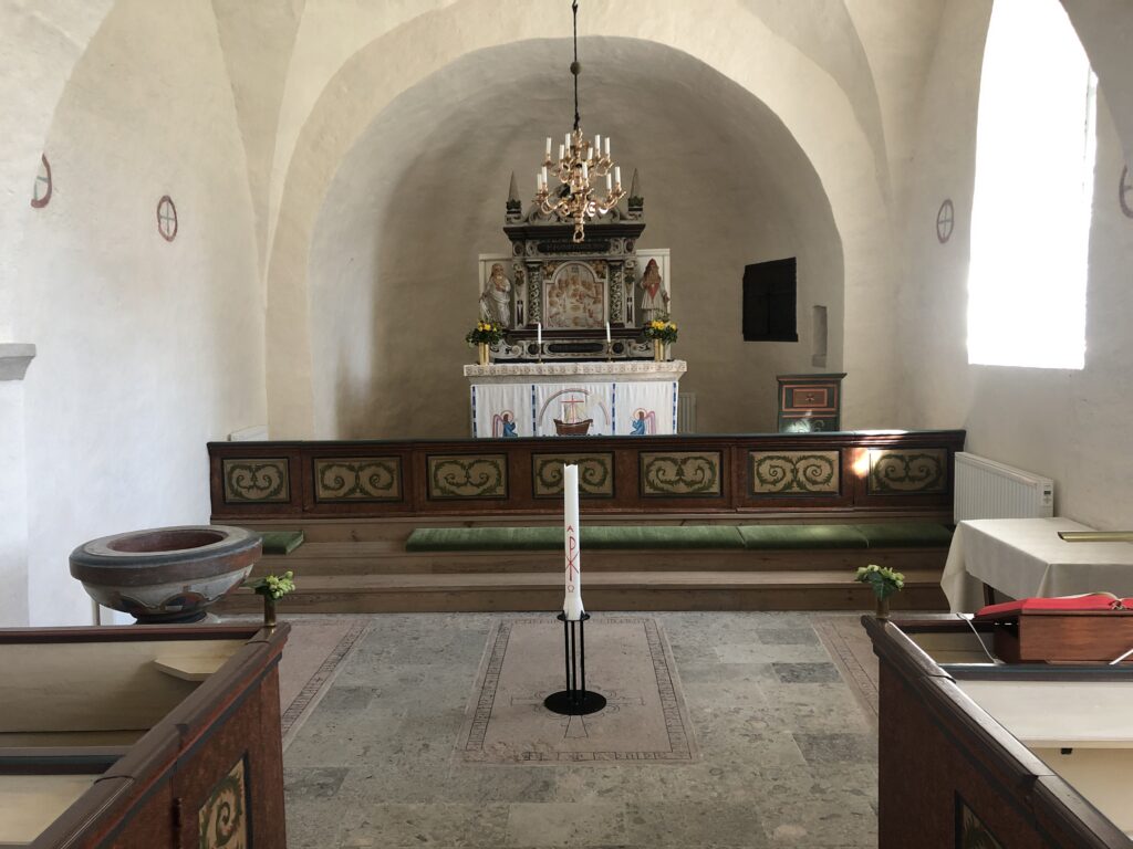 insidan av koret i en kyrka med tre gravhällar liggande i golvet. På den mittersta står ett stort ljus.