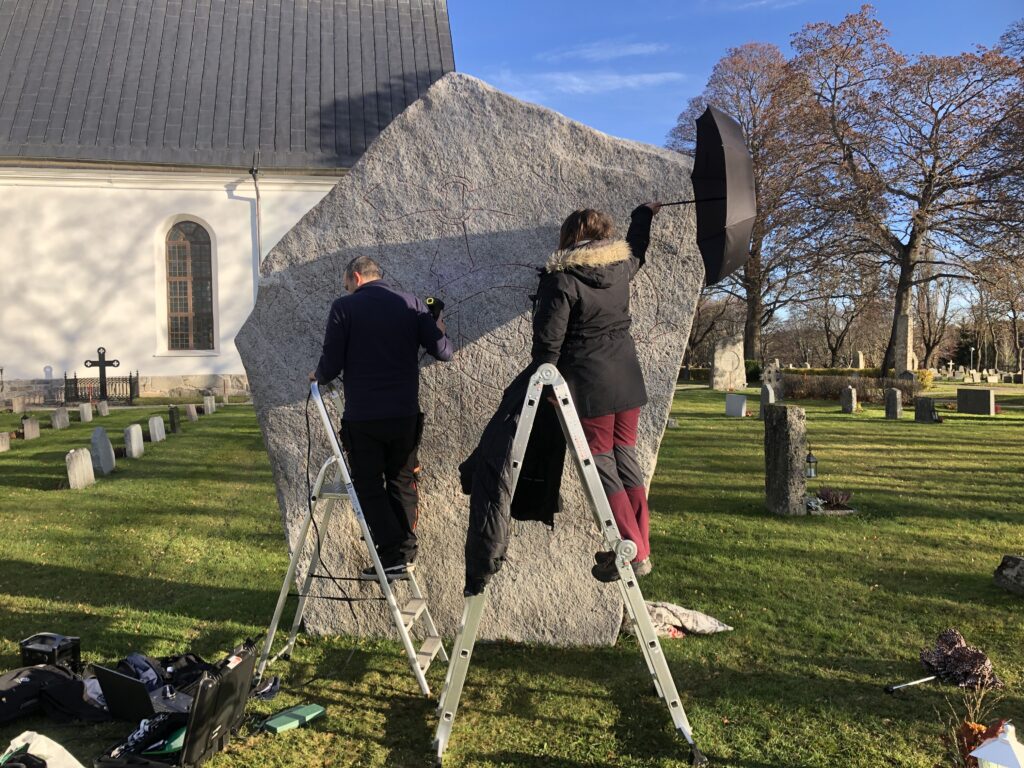 Två personer står på stegar framför en stor runsten. En har en handhållen skanner i handen, den andra skuggar ytan med ett paraply.