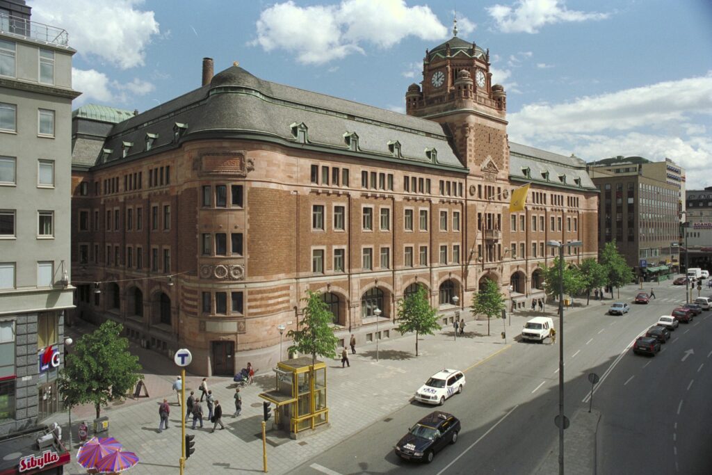 En stor äldre tegelbyggnad med klocktorn, med en gata i förgrunden.