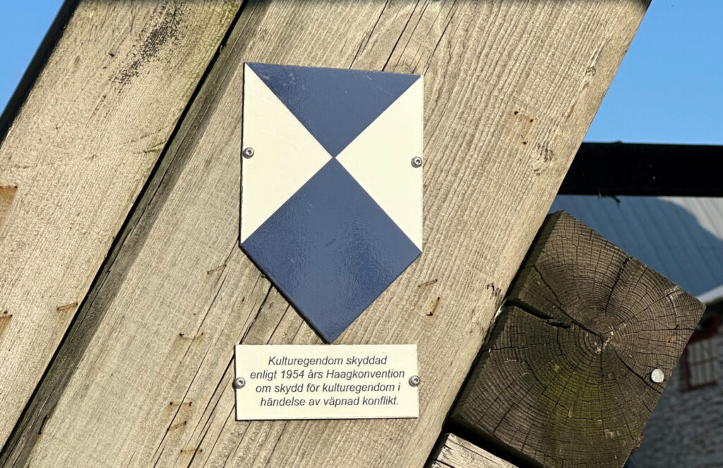 En blåvit skylt. Under skylten står texten "Kulturegendom skyddad enligt 1954 års Haagkonvention..."