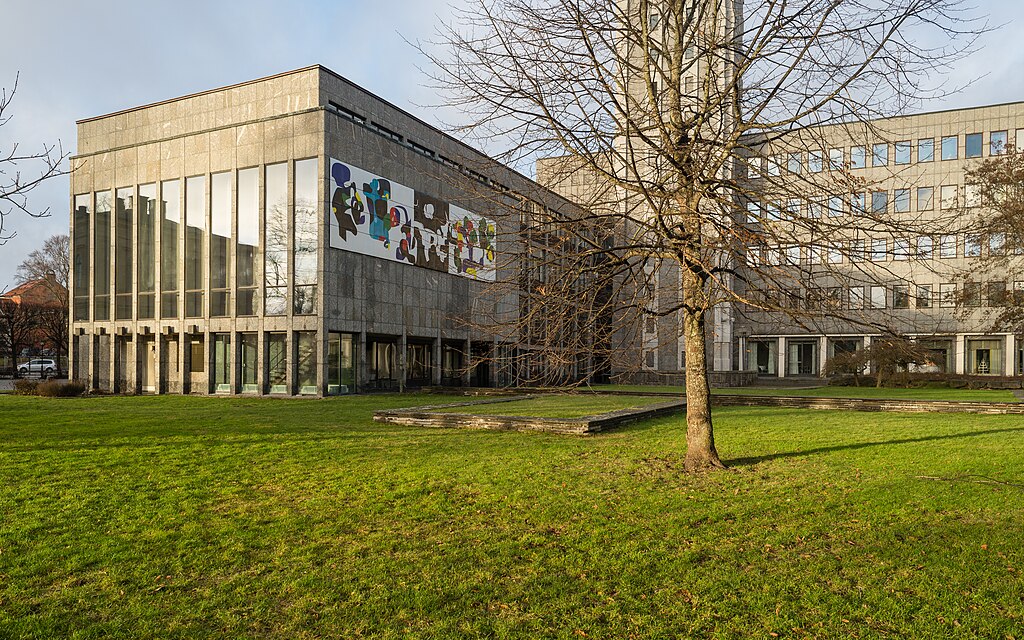 Modernistisk byggnad med stora fönster och betongfasad, prydd med en färgstark muralmålning. Framför byggnaden finns en grön gräsmatta och ett träd utan löv, vilket antyder vintertid.”