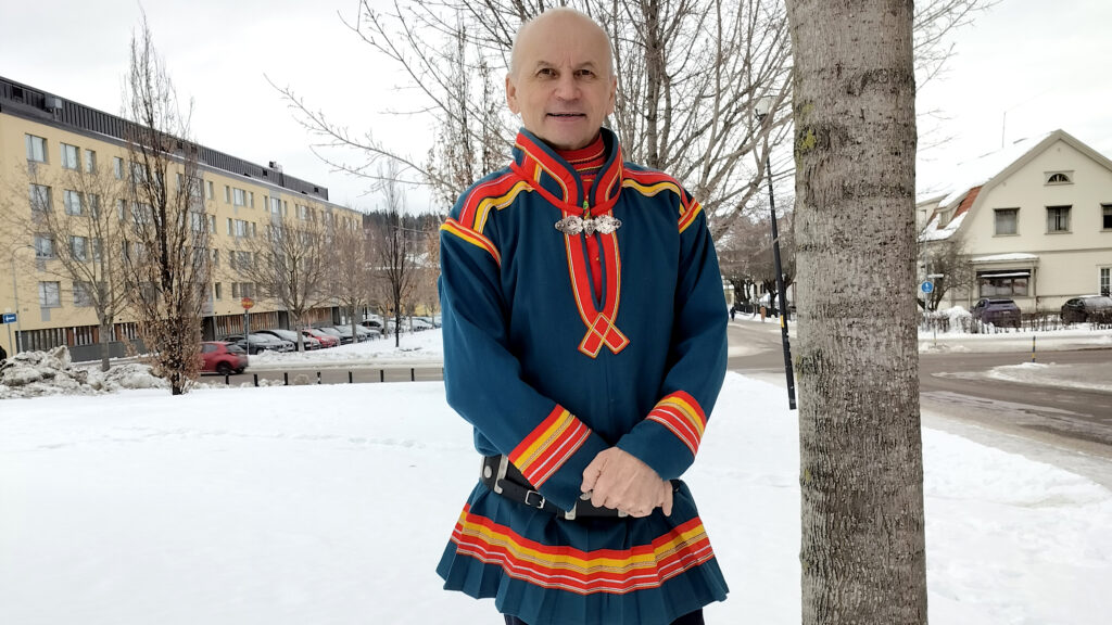 Man med blå samedräkt och färgglada kantband, står framför en avlövad björk, med snö på marken.
