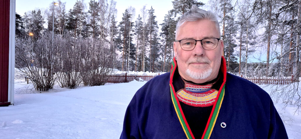 Man med grått hår, skägg och glasögon. Han har en blå samedräkt med färgglada kantband.