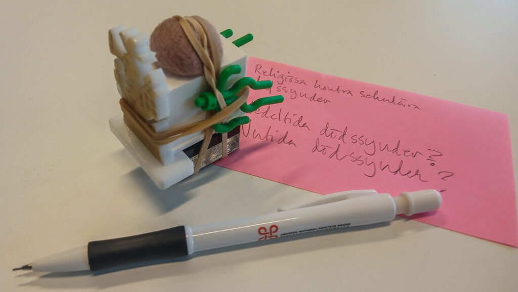 På bilden syns en del av det material som användes vid Riksantikvarieämbetets workshop 2026-02-04. Längst fram ligger en tryckpenna med Riksantikvarieämbetets logotyp. Intill den står en modell av någonting odefinierat, byggd av en 3D-printad kloss, gummisnoddar, legoväxter, en plastfigur och en tovad boll. I bakgrunden syns en rosa postitlapp, med anteckningar i blyerts: ”Religiösa kontra sekulära dödssynder. Medeltida dödssynder? Nutida dödssynder?”
