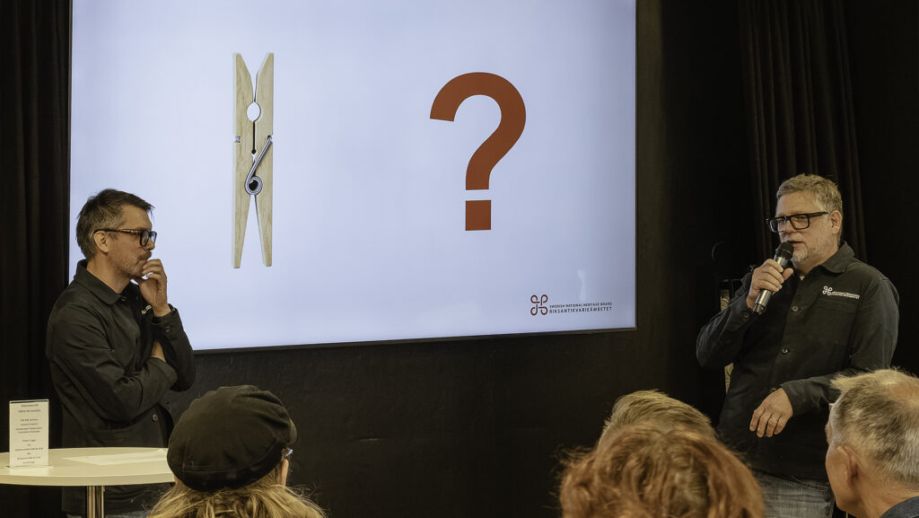På en scen, på en projektorduk, visas en bild av en klädnypa i trä och ett frågetecken. Två män i glasögon och svarta skjortor med Riksantikvarieämbetets logotyp i vitt står på varsin sida om bilden, en med mikrofon i handen